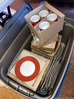 Plastic bin containing stacked boxed ceramic dinnerware items including cups, plates, and bowls all with red rim pattern.