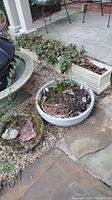 Photo showing a long rectangular cement/resin planter attached to porch steps, a large round gray pot with soil and plants, and smaller pottery pieces nearby.