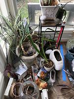 Photo of a corner with multiple potted plants, garden figurines including mushroom shapes, a black metal plant stand holding a white pot and figurines, a large striped metal pot on the floor, and scattered gardening supplies and small pots.