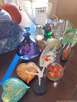 Photo displaying a variety of colorful glass art and figurines including paperweights, a ballerina figurine, and decorative glass pieces arranged on a table.