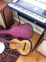 Electronic keyboard mounted on black folding stand above open guitar case on floor, speaker cabinet at left.