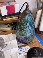 Front view of the colorful teardrop glass pendant lamp with bronze fittings resting on a table, showing its intricate pattern and attached cord.