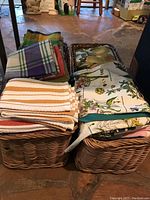Photo showing two wicker baskets filled with assorted kitchen linens in different patterns and colors, including striped and fruit-themed fabrics.