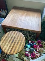 Square wooden table and round pedestal stool beside it on patterned carpet near walls and boxes. Table has scratches and stool shows striped wood pattern with stains.