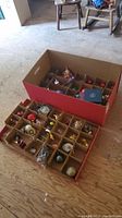 Two cardboard storage boxes filled with various vintage Christmas ornaments including balls, birds, and novelty shapes arranged in compartments.