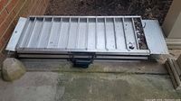 Photo of folded aluminum ramp stacked on the ground next to brick wall and concrete surface.