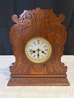 Front view showing ornately carved wooden clock case with brass bezel and Roman numeral clock face.