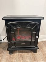 Front view of Duraflame compact electric fireplace showing black metal body, glass front with ember simulation, and control knobs on the side.