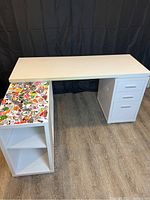 Full view of large white modular desk with three drawers on right and shelving with stickers on left.