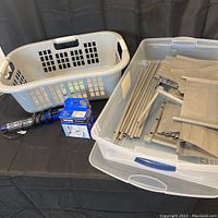 Photos showing grey fabric shoe rack components disassembled in clear rollaway storage bin, white plastic laundry basket, and boxed Moen Banbury chrome robe hook with black Conair hair styling brush on table.