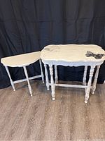 Two antique white painted wooden side tables with distressed finish, photographed against a black backdrop on wooden floor, showing overall shape and size difference.