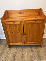 Front view of pine wood cabinet showing double doors and single drawer with metal knobs, warm honey finish, visible wood grain and knots.