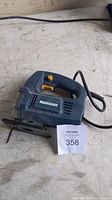 Photo of a blue and black Mastercraft electric jigsaw with attached power cord and visible blade, placed on a workbench with a MaxSold tag number 358.