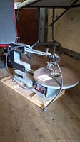 Front side view of the scroll saw showing the saw table, saw arm, and control switches, with the saw mounted on a wooden pallet.