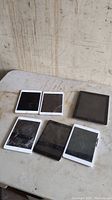Six Apple iPads arranged on a table, four with cracked screens clearly visible, varying bezel colors.