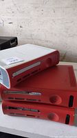 Three Xbox 360 consoles stacked; two red units and one white unit with a red faceplate showing the front panel and disc drive