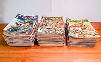 Three stacks of Conan comic magazines laid on wooden surface, showing edges and thickness, confirming quantity and condition.