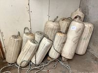 Five large and six small white cylindrical marine buoys arranged in a corner, showing wear and attached rope handles.