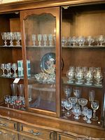 Glassware display inside a wooden cabinet with glass doors including multiple shelves filled with various crystal and glass glasses and a floral plate in the center shelf.