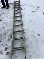 Photo showing the full length of an aluminum extension ladder laid out on snowy ground with visible rungs and parts of a person's legs nearby.