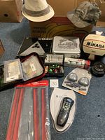 Lot of assorted fishing lures, hooks in plastic bags, boxed games, sports equipment, compass, remote, and hats laid out on floor