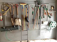 Wide view of pegboard wall with various long-handled garden tools hung including shovels, rakes, hoes, and weed pullers to show inventory.
