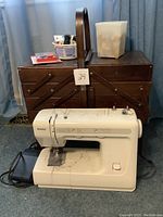 Kenmore sewing machine with foot pedal and power cords arranged in front of closed wooden sewing basket on carpet floor.