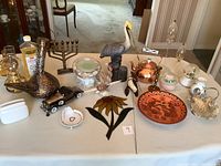 View of all lot items on table including pelican sculpture, menorah, copper kettle, snowglobes, stained glass sunflower, decorative plate, and assorted small items