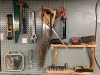 Photo shows a variety of hand tools mounted on a pegboard and wood shelves including saws, hammers, tape measure, socket set, wrenches, level, and hex keys.