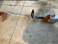 Photo showing entire Stihl gas weed eater laid on concrete floor, visible curved shaft, and orange string guard at the cutting end.