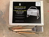 Box label showing the brand PC tabletop propane gas grill with 11,000 BTU rating and image of the stainless steel grill. Box also states 'canister not included'.