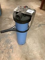 Full view of blue water filter housing showing the black cap, mounting bracket, and wrench attached around the base.