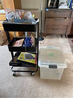 Image of black rolling cart with artist supplies like chalk, markers, crayons, paint brushes, gel pens, and stickers in trays along with a large clear plastic container next to it