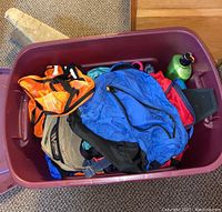 Plastic storage bin filled with various backpacks and bags including an orange black bag, blue backpack, gray backpack, and green metal water bottle.