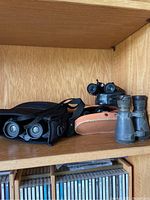 Three sets of binoculars on wooden shelf with two carrying cases: a smaller black case, a beige leather case, and a vintage pair without visible case.