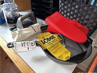 Photo showing two Lodge cast iron pans, one round skillet and one rectangular grill pan with tags, along with a red silicone oven mitt and a white fabric oven mitt.