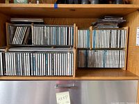 Wooden rack shelving filled with approximately 50+ assorted music CDs in jewel cases, arranged with spines out for title visibility.