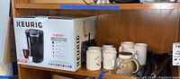 Photo of Keurig K-Select coffee maker in box placed on wooden shelf with ceramic mugs beside it.