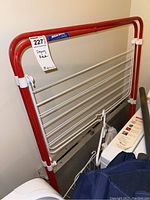 Front view of foldable metal drying rack with red frame and white rods leaning against a wall.