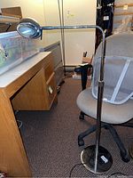 Full view of the chrome floor lamp showing its height, base, and adjustable arm with head, positioned beside an office chair and desk.