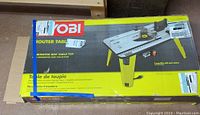 Boxed Ryobi router table with yellow foldable legs shown on packaging.
