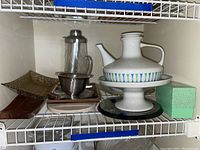 Shelf with various serveware items including Contempri coffee pot, glass cake plate, stainless steel serving pieces, egg cups, and green patterned box.