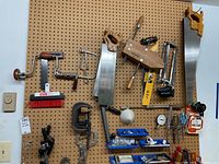 General overview of several hand tools and hardware items hanging on a pegboard, including saws, clamps, a hand drill, brush, and organizer bins.