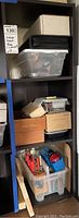 Full view of three shelves holding plastic storage bins, wooden and cardboard boxes filled with various hand tools including X-Acto knives and blades, measuring tape and assorted cutters.