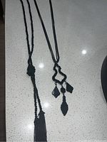 Photo showing two black necklaces; one has beads, a knot and tassel, the other has a cord and three geometric black pendants.