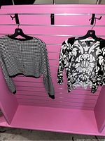 Two black and white patterned women's tops hanging on black hangers against a pink background; left is houndstooth cropped sweater, right is floral cardigan.