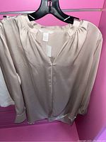 Beige satin-like blouse with a gathered neckline and long sleeves hung on a black hanger in front of pink background.