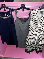 Three women's dresses hanging on black hangers against pink backdrop: navy dress with beaded neckline, gray dress with sweetheart neckline, white dress with black stripes and green accents.