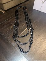 Photo of a black 3-tier beaded necklace laid flat on dark wood surface, showing the layered design with faceted and round beads and chain extension.