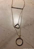 Photo of a 3 tier gold-tone chain necklace showing a heart-shaped pendant with dark stones and a circular pendant with multi-colored stones, laid flat on a surface.